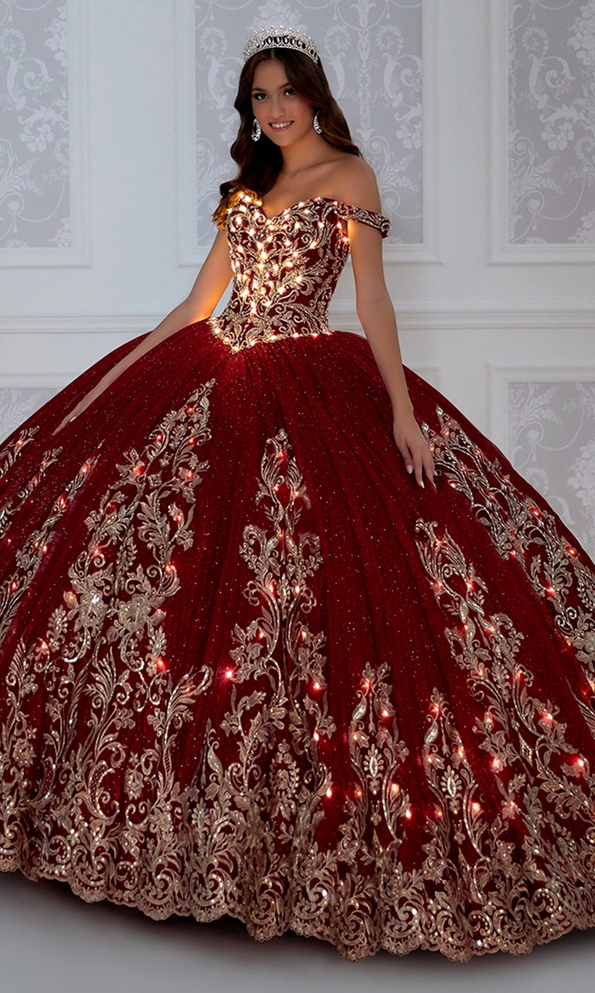 A young woman in a tiara wears the PR12264 Princesa Quince Dress with gold floral embroidery, glitter tulle skirt, and glowing lights, standing against a white patterned wall.